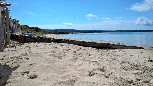 Ein Sandstrand mit einer Holzpromenade, die zu einem Gewässer führt.