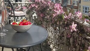 Ein runder Tisch mit einer Schüssel Erdbeeren und einer Zeitschrift, ein Stuhl sowie im Hintergrund der Blick auf Gebäude und das Meer.