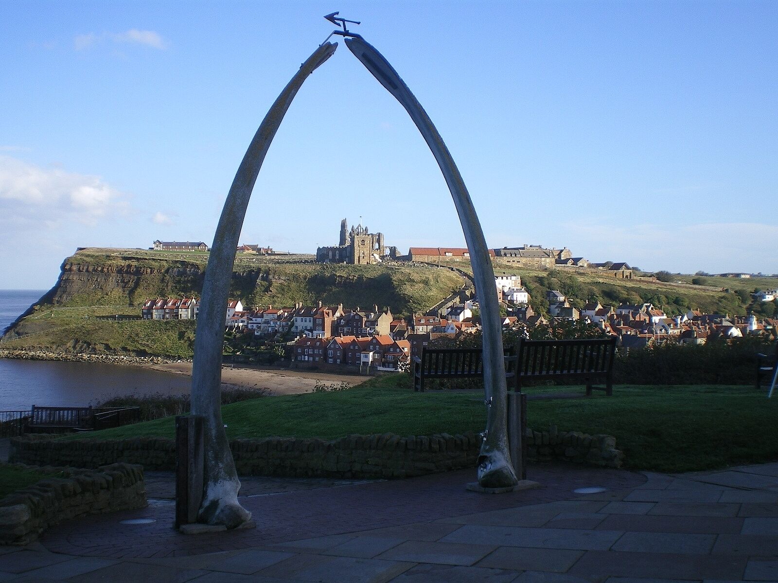 A coastal town with a prominent church on a hill, a stone archway in the foreground, and a bench.