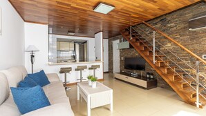 Un salon moderne avec un plafond en bois, un canapé avec des coussins bleus, une table basse blanche, un coin cuisine et un escalier en bois.