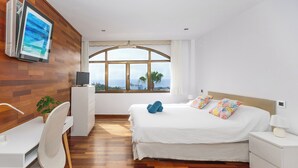Une chambre d’hôtel moderne avec un mur en bois, une grande fenêtre offrant une belle vue, un lit avec une literie blanche et des oreillers colorés, un bureau avec une chaise et un téléviseur fixé au mur.