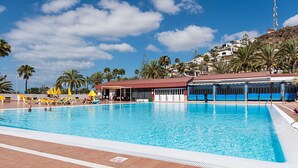 Une piscine à l’eau d’un bleu limpide, entourée de chaises longues et de parasols, avec un bâtiment et des palmiers en arrière-plan.