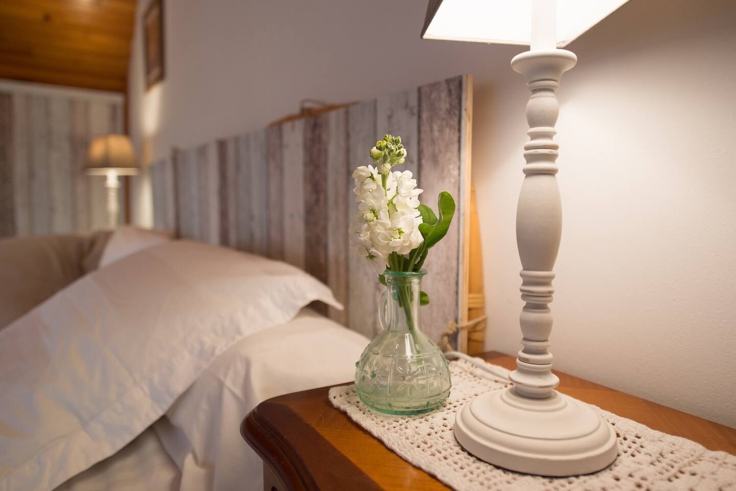Ein ordentlich bezogenes Bett mit weißen Leinentüchern, eine kleine Glasvase mit weißen Blumen auf einem Nachttisch aus Holz und eine weiße Tischlampe.