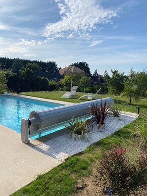 Ein Schwimmbecken mit modernem Filtersystem, umgeben von einem angelegten Garten mit verschiedenen Pflanzen und einem Reetdachschirm.