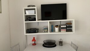 A neatly organized room with a white table, chairs, a TV on a shelf, and various items on the shelves including books, a lamp, and a basket. - Spacious, modern ground floor apartment, private patio and garden, mosquito nets (Calabria)
