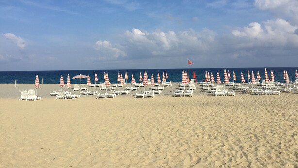 A beach with rows of white lounge chairs and red and white striped umbrellas. - Spacious, modern ground floor apartment, private patio and garden, mosquito nets (Calabria)