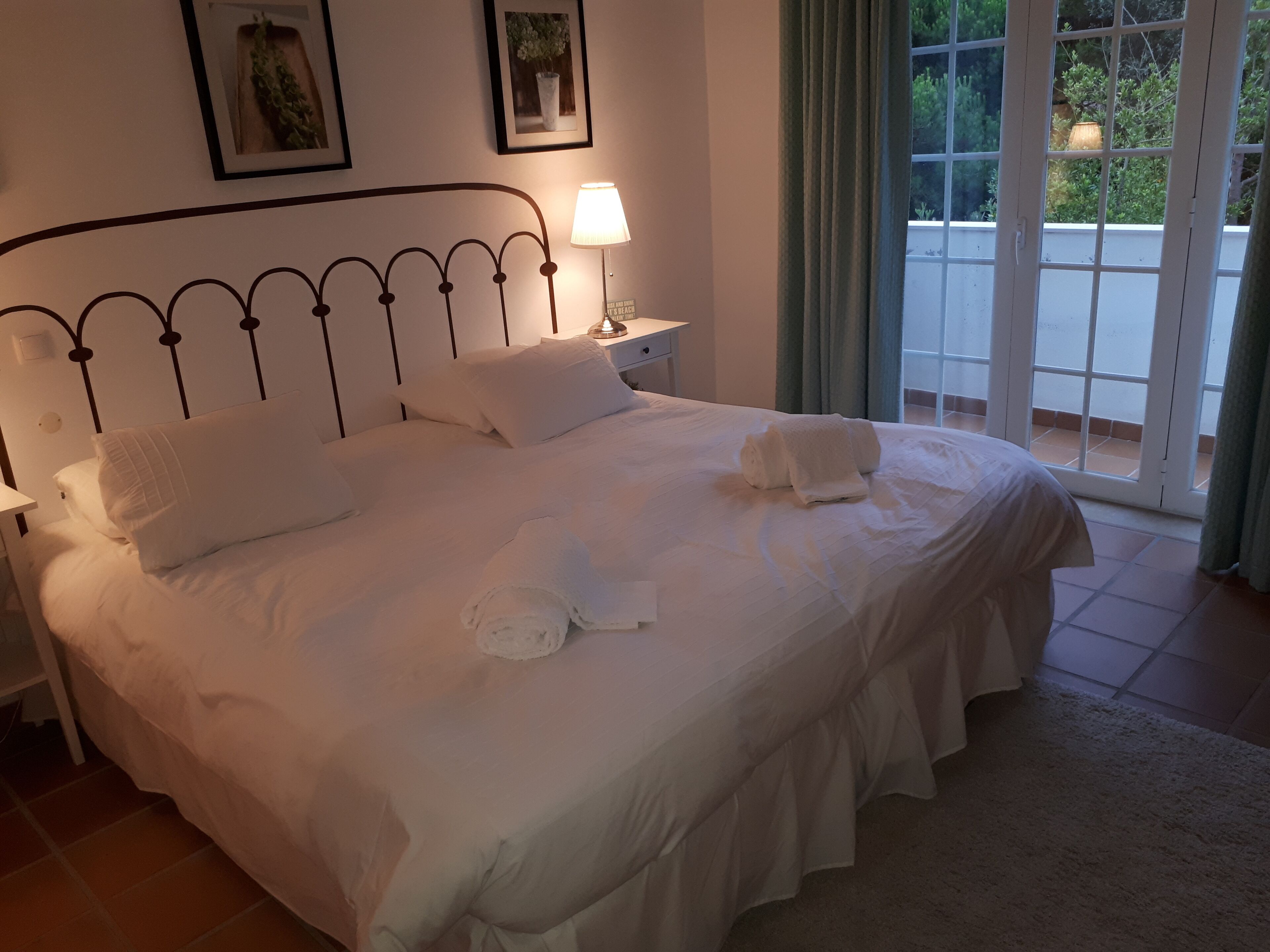Una cama bien hecha con sábanas y toallas blancas, una lámpara de noche y vistas a un balcón a través de una puerta de cristal.