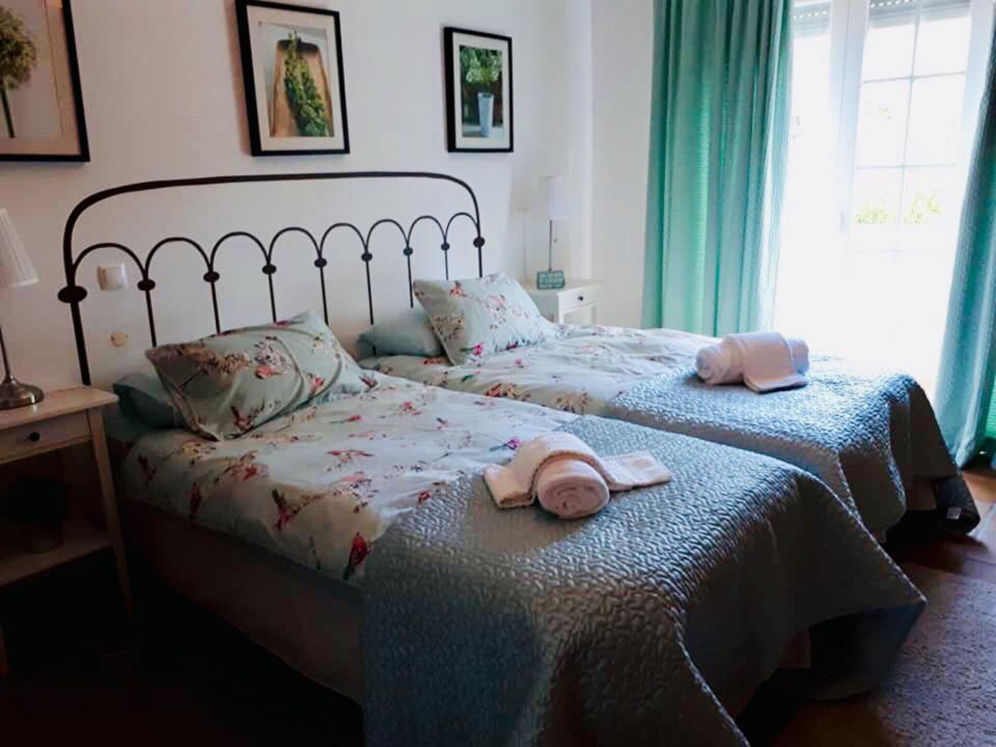 Una cama doble con ropa de cama floral y cabecera de metal. Hay dos mesitas de noche, una con una lámpara y la otra con una pequeña planta. Se ve una ventana con cortinas.
