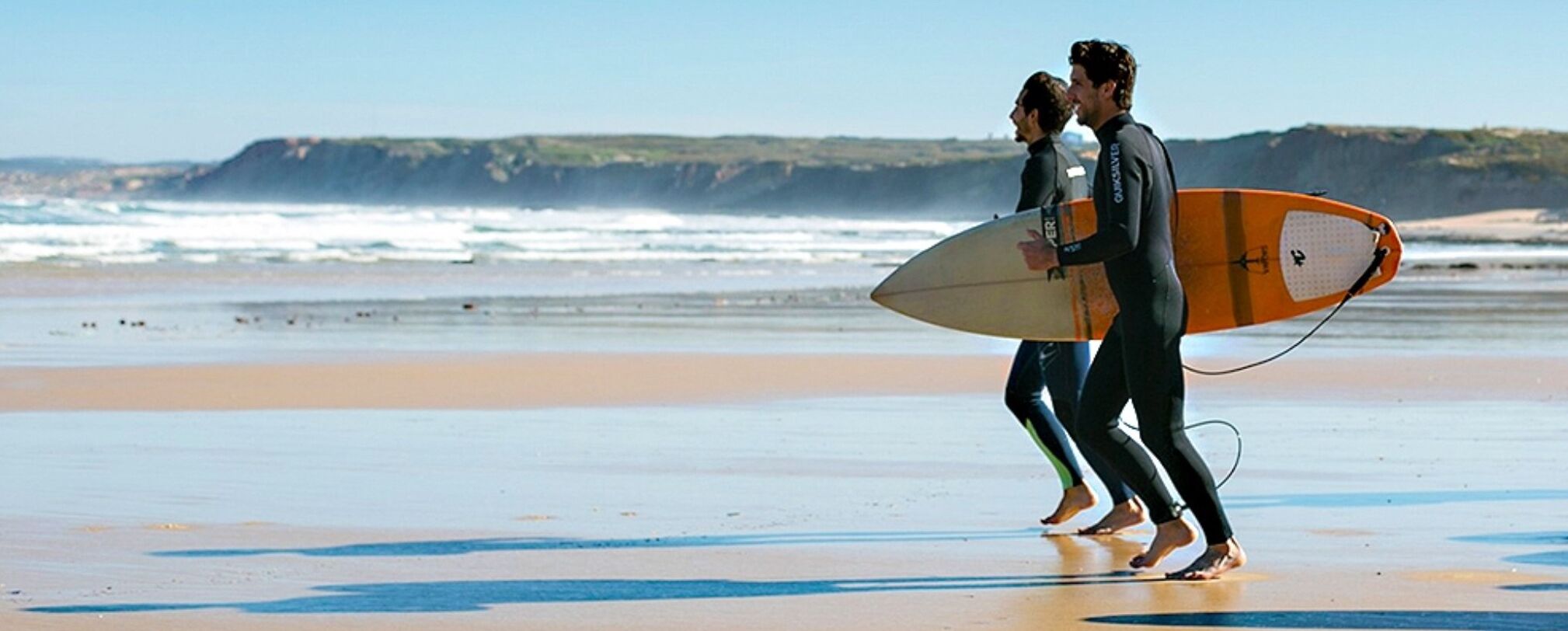 Two people walking on a beach with surfboards.