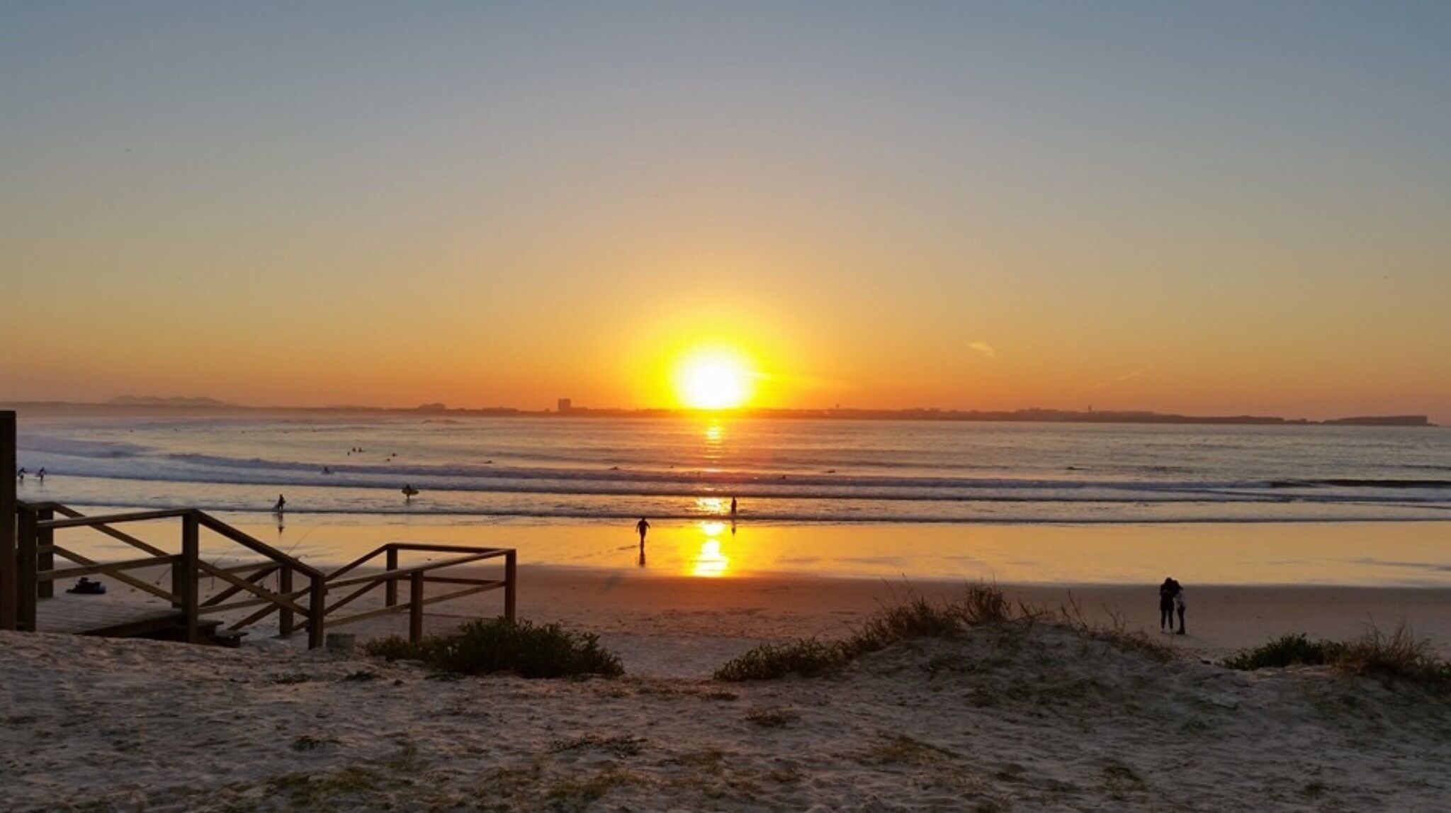A beach with a wooden walkway leading to the water, waves, and a sunset.