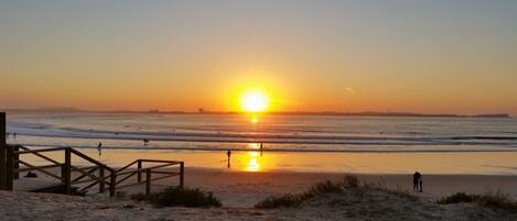 A beach with a wooden walkway leading to the water, waves, and a sunset.
