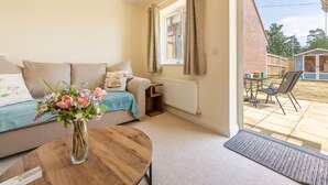 Un salon chaleureux avec un canapé, une table basse et un vase de fleurs. Il y a une fenêtre avec des rideaux, un radiateur et une vue sur le jardin, qui comprend une terrasse et du mobilier d’extérieur.