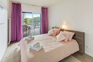 A bedroom with a large bed, a sliding glass door to a balcony, and a table with chairs outside.
