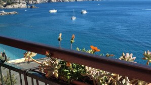 Vista costera con barcos en el agua, montañas al fondo y un balcón con plantas en macetas.