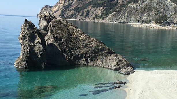 Un promontorio rocoso en aguas transparentes, con una playa arenosa y montañas al fondo.