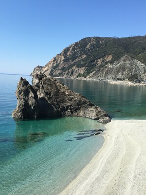 A coastal landscape with a rocky outcrop in clear water, a sandy beach, and a mountainous backdrop.