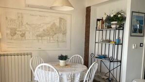 Un comedor con una mesa redonda y sillas blancas, un radiador, un estante en la pared con libros y adornos, y una lámpara colgante.