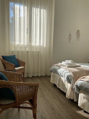 Une pièce comprenant un lit, deux fauteuils en osier, une fenêtre avec des rideaux légers et un élément décoratif fixé au mur.