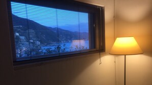 Una habitación con ventana que da a un paisaje montañoso, una lámpara de noche y una cama con cubrecama blanco.