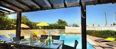 An outdoor pool area with a table and chairs, umbrellas, and a view of the sky and distant structures.
