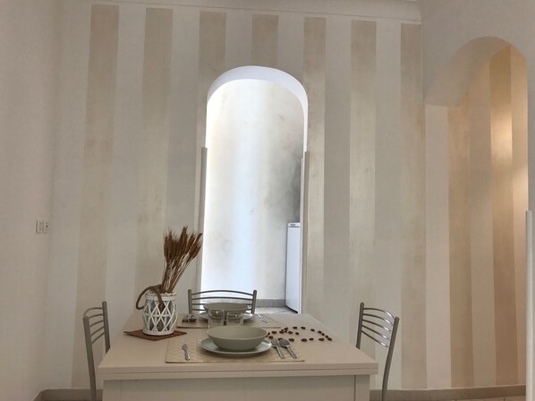 Un coin repas avec une table dressée pour deux personnes, un vase blanc contenant des plantes séchées et un purificateur d’air blanc.