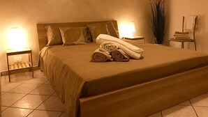 Ein ordentlich bezogenes Bett mit aufgerollten Handtüchern, zwei Nachttischlampen, ein Stuhl mit Handtuch und eine hohe Pflanze neben dem Bett.