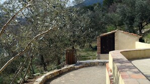Um caminho de pedra leva a um edifício com telhado em cerâmica, cercado por oliveiras e uma paisagem montanhosa.
