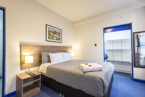 Una camera d'albergo con un letto, un comodino con una lampada, un quadro appeso al muro e un tappeto blu.