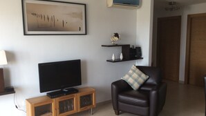 A living room with a flat-screen TV, a leather armchair, a coffee table, a wall-mounted shelf with decorative items, and a framed artwork. - Hacienda Riquelme Polaris World Golf / Holiday Resort, Sucina, Spain  (Murcia)