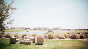 A grassy outdoor area with wicker furniture and a view of a lake and buildings in the background. - Hacienda Riquelme Polaris World Golf / Holiday Resort, Sucina, Spain  (Murcia)