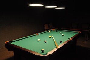 Una mesa de billar con bolas y taco, rodeada de sillas e iluminación colgante.