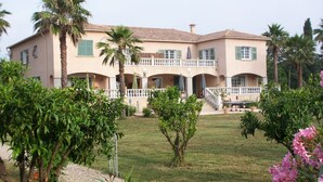 A two-story building with a balcony, surrounded by a well-maintained garden with palm trees and flowering plants.