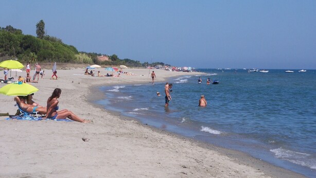 A beach with people sunbathing and swimming, umbrellas, and boats in the water.