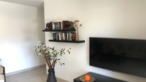 Ett modernt vardagsrum med en platt-TV, en bokhylla med böcker och dekorativa föremål, en vas med blommor och ett litet orangefärgat ljus på ett svart ljusställ.