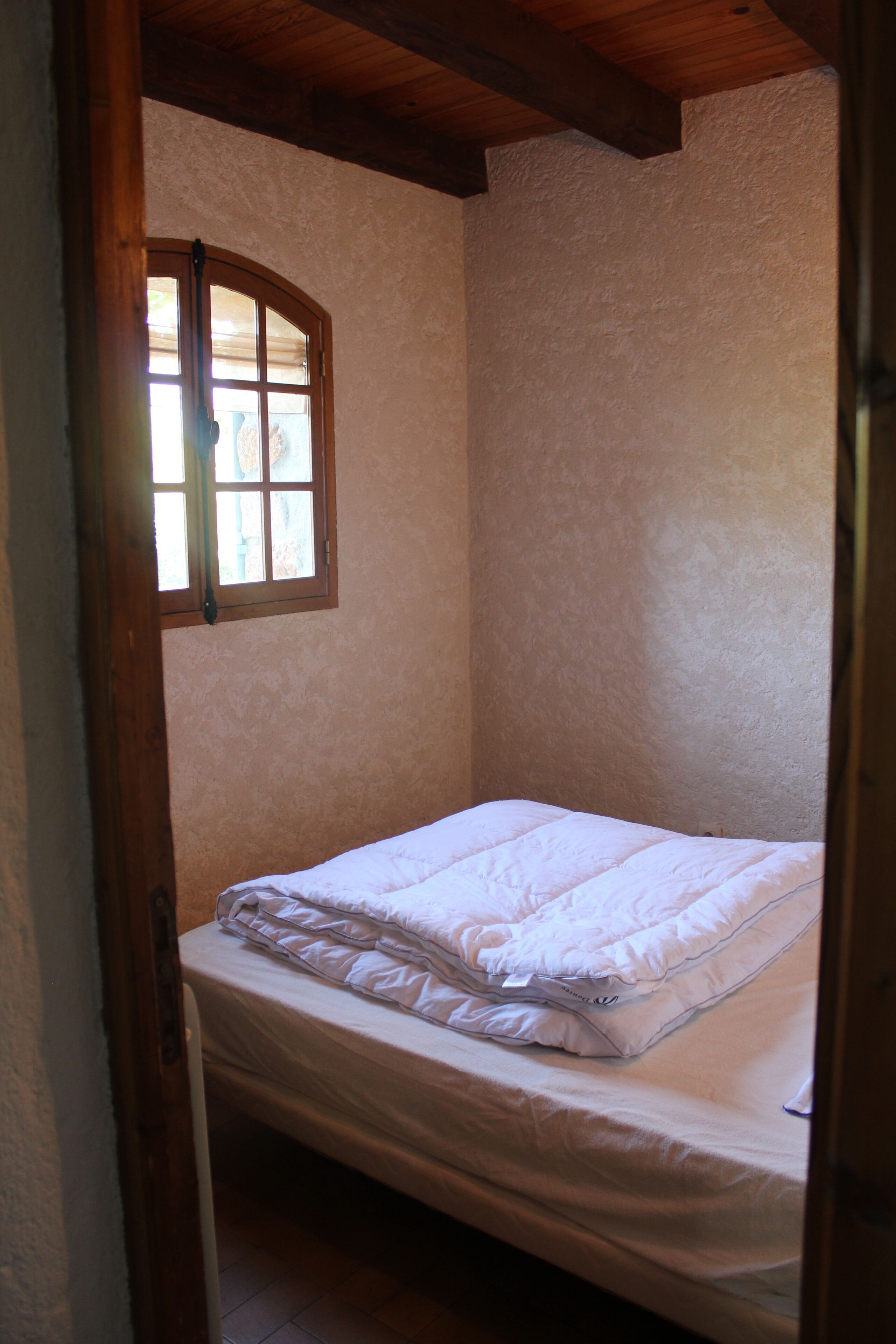 Une petite pièce étroite, avec un lit simple, une fenêtre et une poutre en bois au plafond.