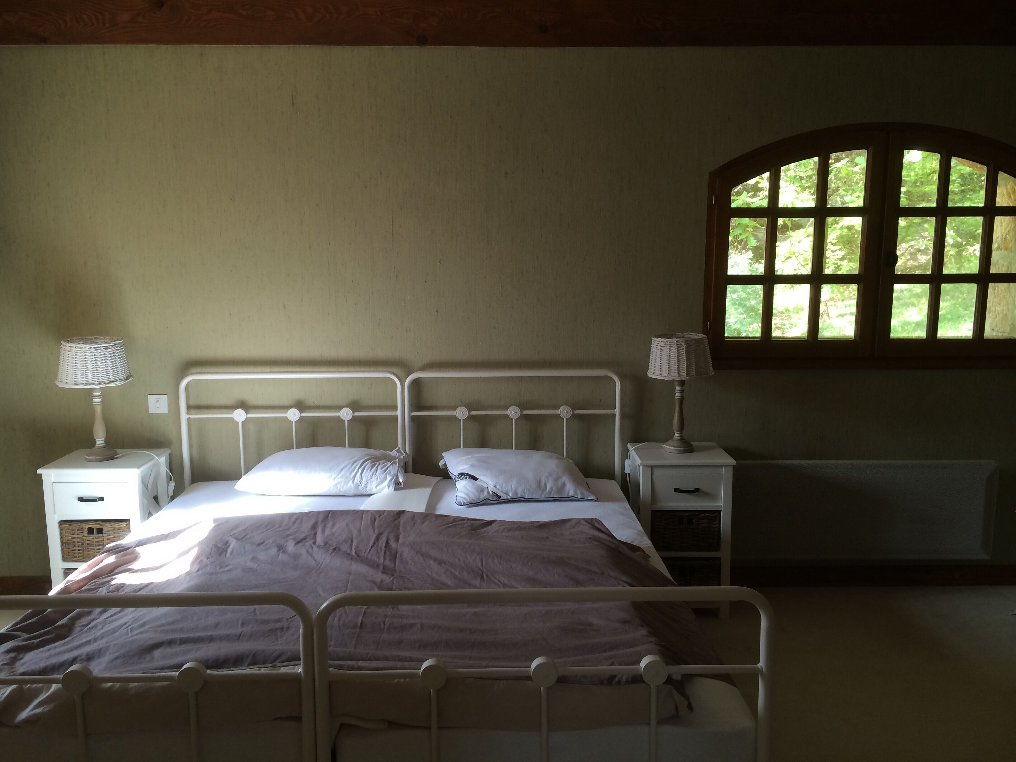 Une chambre avec un lit simple, un cadre en métal, deux tables de chevet, une lampe sur chacune, et une fenêtre donnant sur un paysage verdoyant.