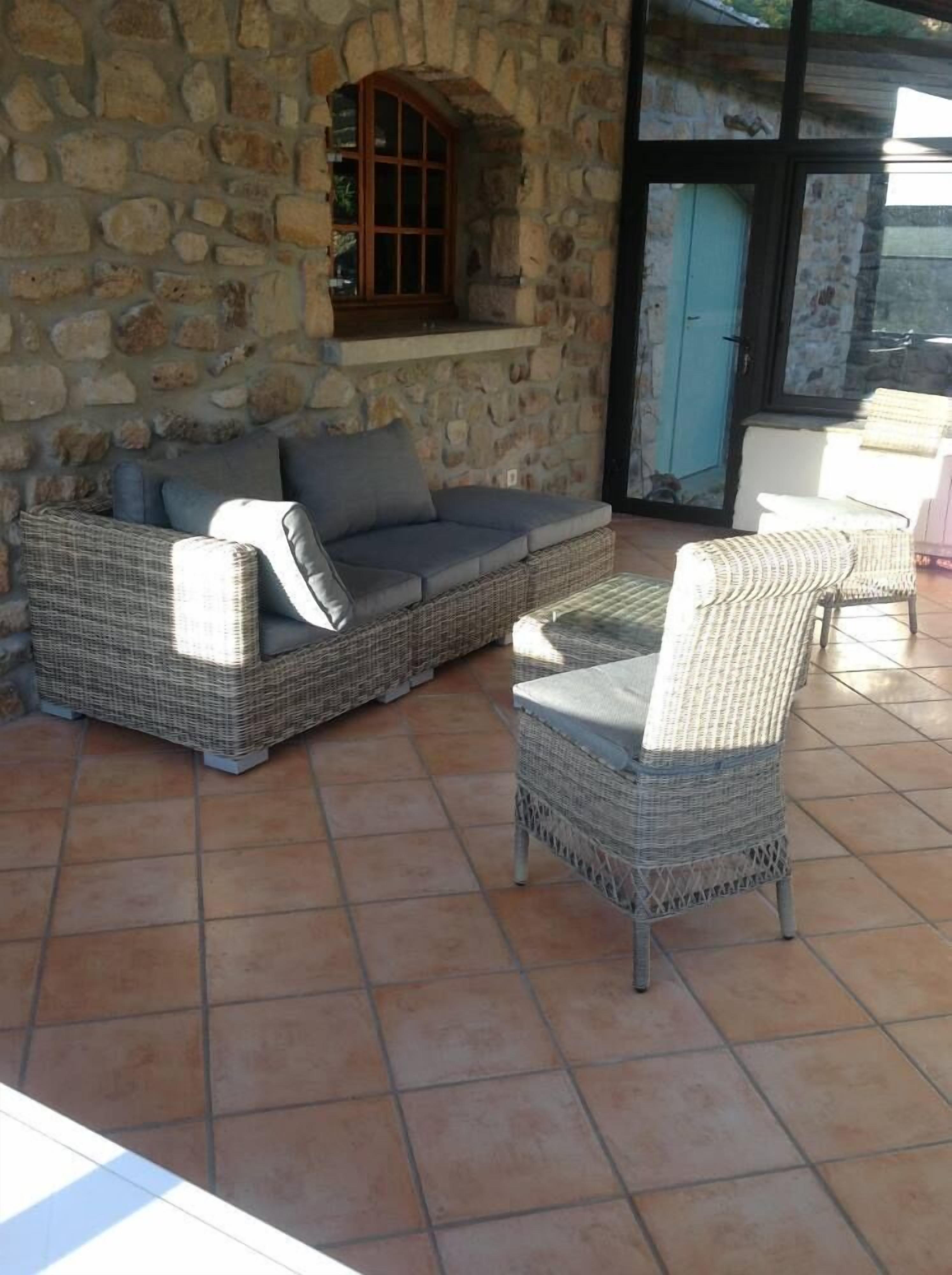 Un patio con paredes de piedra, un sofá y sillones de mimbre, suelo de baldosas y una puerta con marco azul.