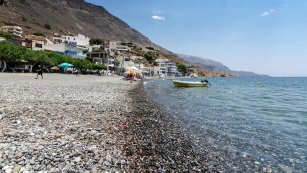 A pebble beach with buildings on a hillside, a boat on the water, and a clear sky. - sunset home crete 1  (heraklion)