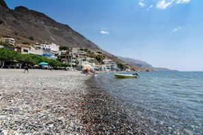 Ein Kiesstrand mit Gebäuden an einem Hang, ein Boot auf dem Wasser und ein klarer Himmel.