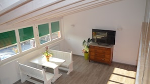 Ein Zimmer mit einem weißen Tisch und einer Bank, einem Fernseher auf einer Holzkommode und einer Topfpflanze.