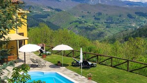 A pool area with lounge chairs and umbrellas, surrounded by a wooden fence, overlooking a mountainous landscape.