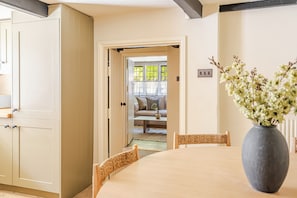 A dining area with a round table, chairs, a vase with flowers, and a view into a living room with a sofa and a window.