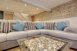A modern living room with a beige leather sofa, striped and blue cushions, and a glass coffee table with pebbles. - Owlets at Mulberry Barn, Heacham, Norfolk (King's Lynn)