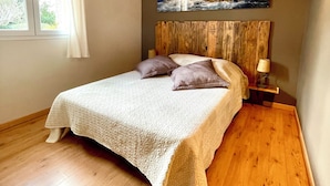 Un dormitorio con techo de madera, una cama con colcha blanca, dos almohadas, una mesita de noche con lámpara y un cuadro de un paisaje rocoso en la pared.