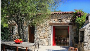 Un edificio de piedra con una puerta roja, una mesa de madera con un frutero y un perro afuera.