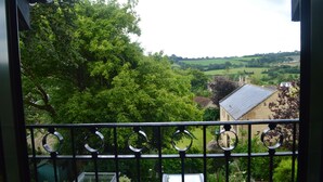 Vue depuis un balcon doté d’une rambarde en métal, donnant sur un paysage verdoyant avec des arbres et un bâtiment surmonté d’un toit en tuiles.