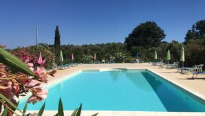 A rectangular swimming pool with a tiled deck, surrounded by lounge chairs and umbrellas, with trees and a clear sky in the background. - Tuscany, an agrotourism apartment between sea and countryside. (Cecina)