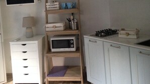 A small kitchen with white cabinets, a microwave, a gas stove, and a wooden shelf with books and cups. - Costa Rei chalet 20 meters from the beach (Costa Rei)
