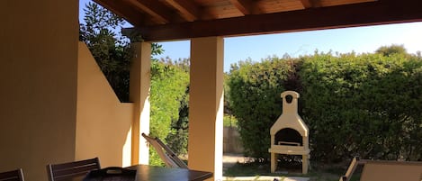 Un patio al aire libre con mesa y sillas, techo de madera y un horno de pizza al fondo.