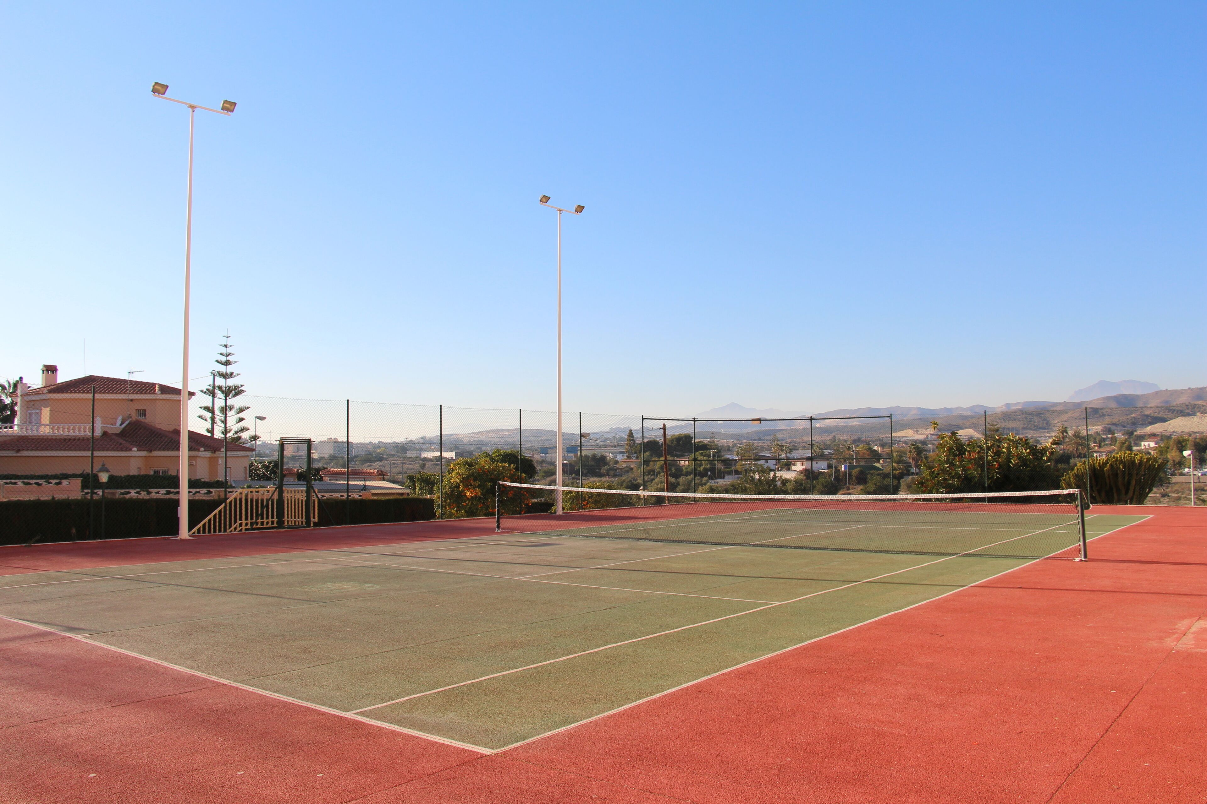 A tennis court with a net, surrounded by a fence, and a clear sky in the background.